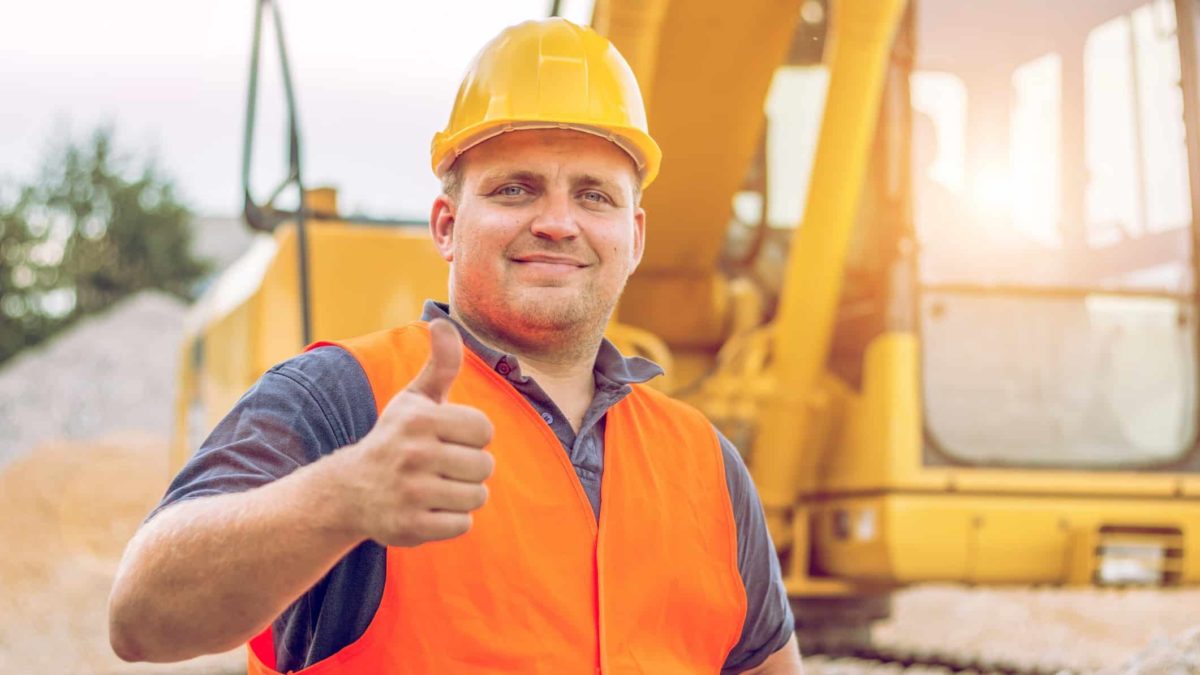 A man in a hard hat and high visibility vest holds his thumb up in a gesture of confidence with heavy moving equipment in the background as on a mine site as the Chalice Mining share price rises today.