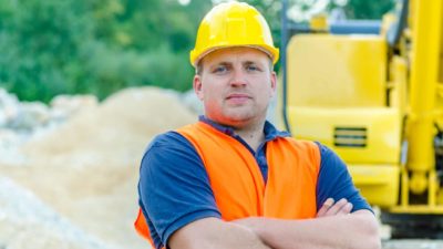 a man wearing a hard hat and a high visibility vest stands with his arms crossed in front of heavy equipment at a mine site.