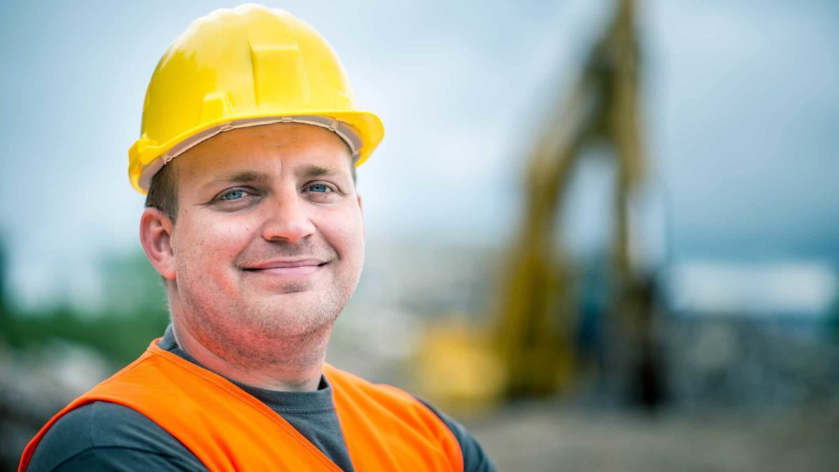a man in a hard hat and high visibility vest smiles as he stands in the foreground of heavy mining equipment on a mine site.