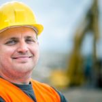 a man in a hard hat and high visibility vest smiles as he stands in the foreground of heavy mining equipment on a mine site.
