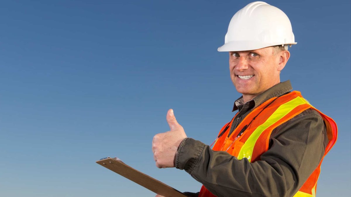 A man in a hard hat gives a thumbs up as he holds a clipboard in one hand against a blue sky background.
