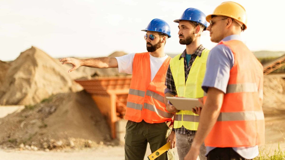 A group of three men in hard hats and high visibility vests stand together at a mine site while one points and the others look on with piles of dirt and mining equipment in the background.