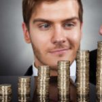 A man with a wry smile on his face is shown close up behind ascending piles of coins as he places another coin on top of the tallest stack representing rising dividends