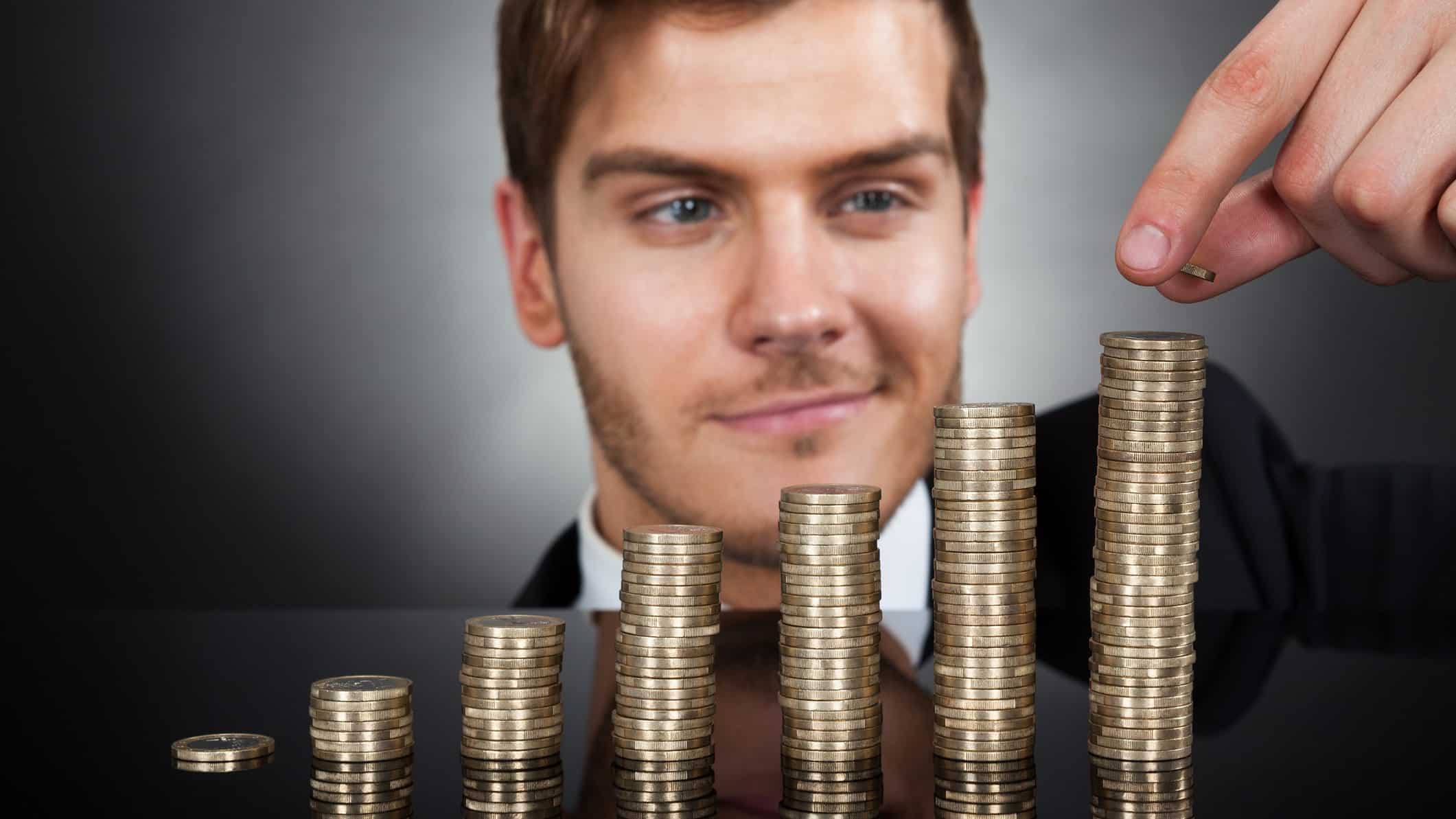 a man with a wry smile is behind ascending piles of coins as he places another coin on top of the tallest stack.