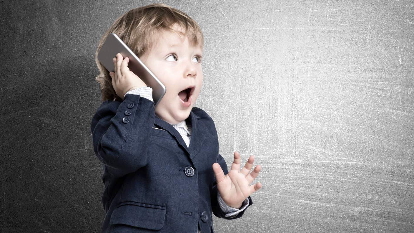 Cute little child is talking on his smartphone while standing in his business suit near a concrete wall.