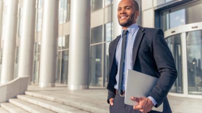 a man in a business suit and carrying a laptop stands smiling with hand in pocket outside a large office building in a city environment.