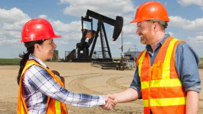 Two workers shake hands in front of an oil rig on the successful completion of a deal.