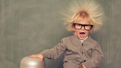 a young child wearing a cardigan and thick black glasses places his hand on a nearly rounded object and his hair lifts at right angles to his head thanks to static electricity.
