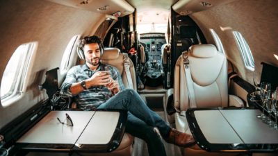 A young cool man sits in a private jet wearing headphones and casual clothing.