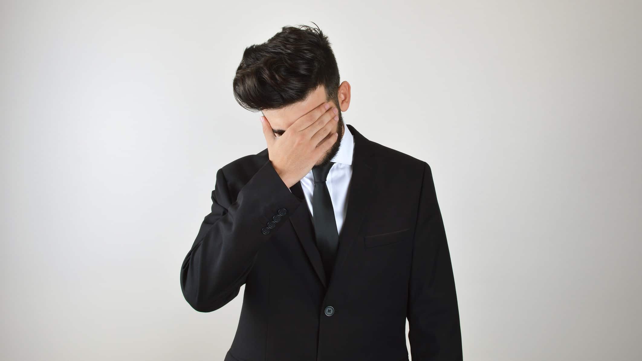 a business man in a suit holds his hand over his eyes as he bows his head in a defeated post suggesting regret and remorse.