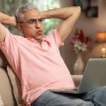 An older man wearing glasses and a pink shirt sits back on his lounge with his hands behind his head and blowing air out of his cheeks.