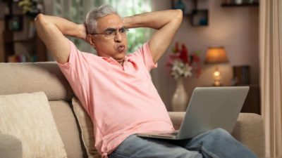 An older man wearing glasses and a pink shirt sits back on his lounge with his hands behind his head and blowing air out of his cheeks.