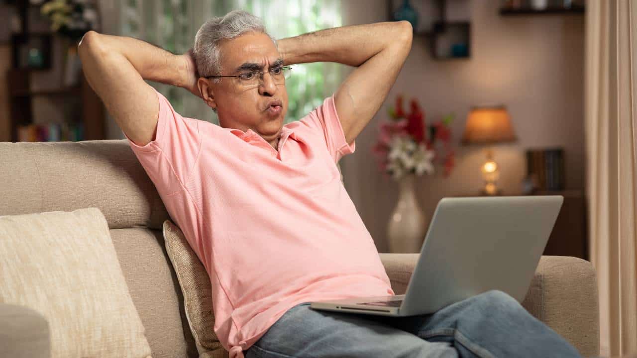 An older man wearing glasses and a pink shirt sits back on his lounge with his hands behind his head and blowing air out of his cheeks as he reads about the Crown share price and anticipated AUSTRAC fines on his laptop