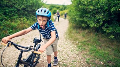 A young boy pushes his bicycle uphill on a rocky road. He is wearing a helmet and has his tongue hanging out as though he is making a face to show how exhausted he is.