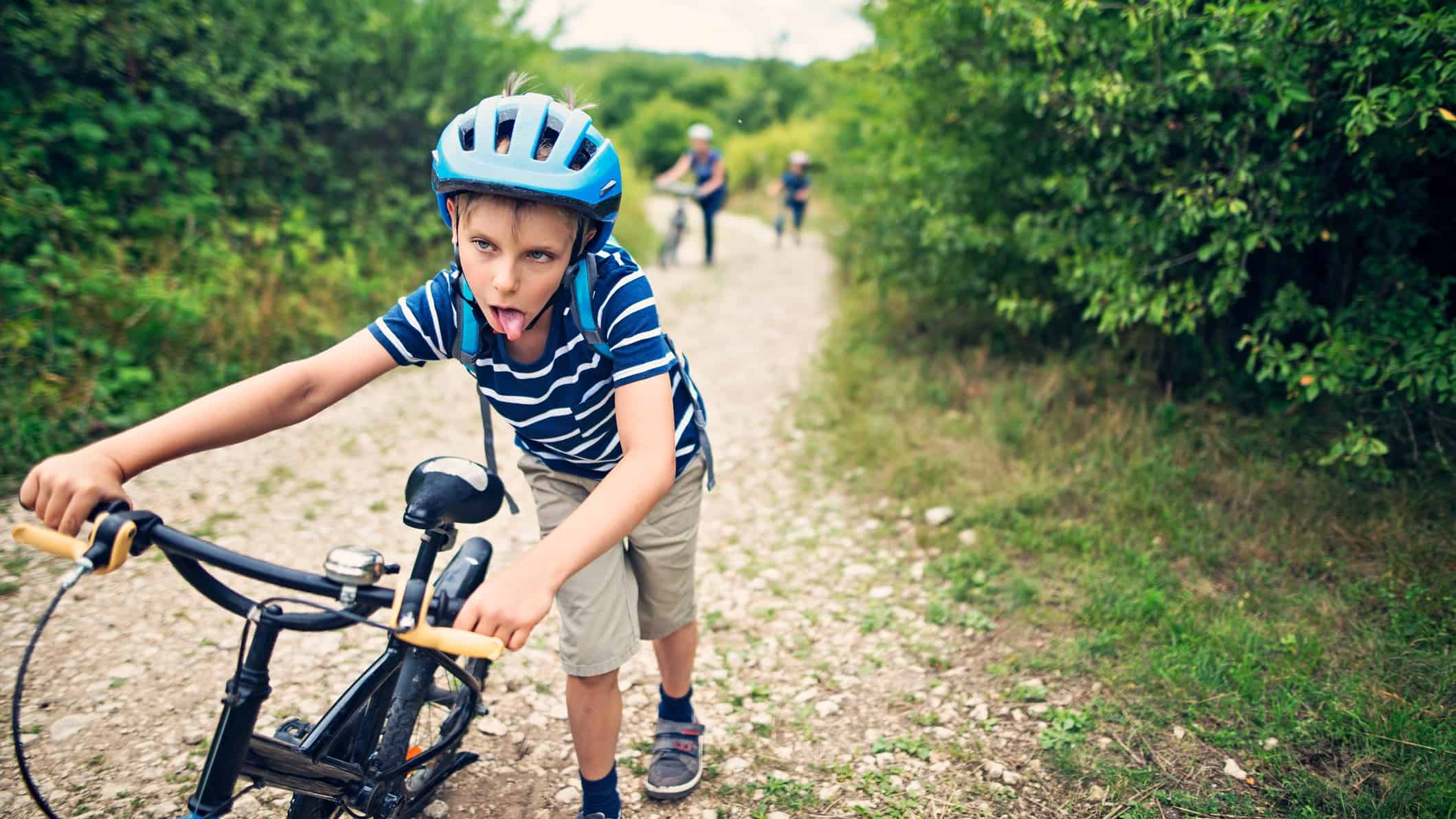 a young boy pushes his bicycle uphill on a rocky road. He is wearing a helmet and has his tongue hanging out as though he is making a face to show how exhausted he is.