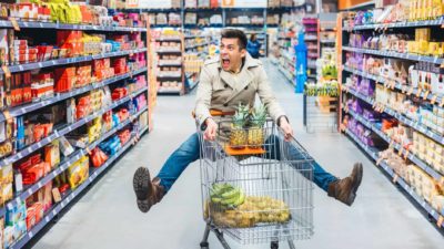 A man in a supermarket strikes an unlikely pose while pushing a trolley, lifting both legs sideways off the ground and looking mildly rattled with a wide-mouthed expression.