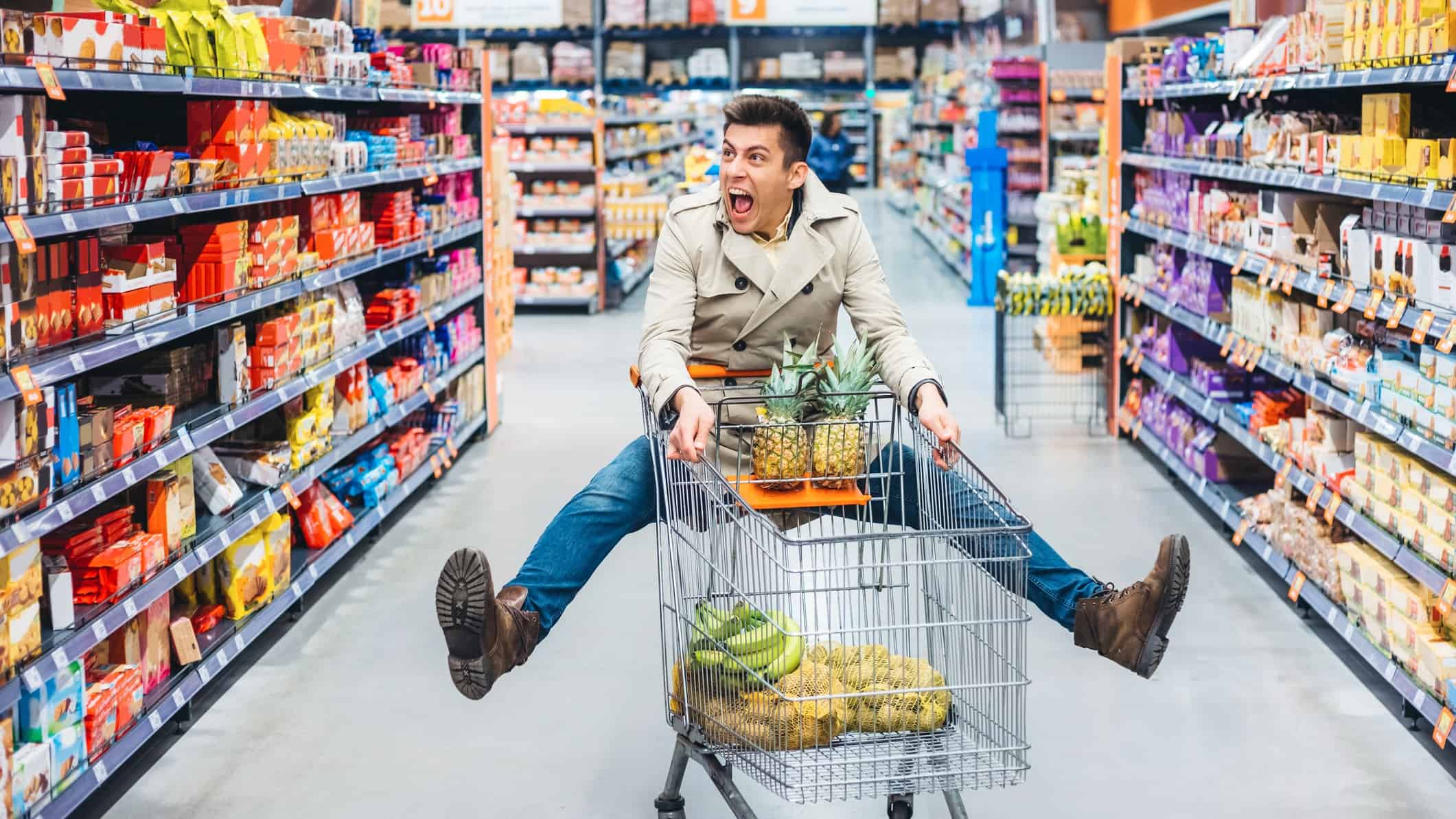 a man in a supermarket strikes an unlikely pose while pushing a trolley, lifting both legs sideways off the ground and looking mildly rattled with a wide-mouthed expression.