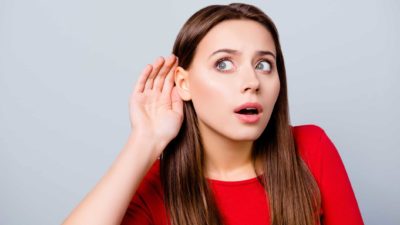 a young woman holds her hand to her ear and leans sideways as if to listen to something that's surprising her as her eyes and her mouth are wide open.