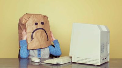 Kid with a brown paper bag on his head which has a sad face on it sits in front of an old style computer representing falling ASX 200 tech shares today.