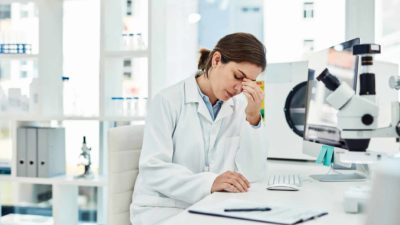 A disappointed lab researcher sits in her lab looking at her clipboard with her hand to her face as she worries about the Imugene share price today