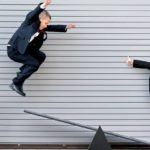 One young boy jumps off a step ladder and is captured mid-air about to land on a see-saw where his friend is standing with a wide smile on his face looking at the camera and holding his thumbs up as though he is excited for the ride to come. Both boys are wearing business suits.
