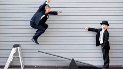 One young boy jumps off a step ladder and is captured mid-air about to land on a see-saw where his friend is standing with a wide smile on his face looking at the camera and holding his thumbs up as though he is excited for the ride to come. Both boys are wearing business suits.