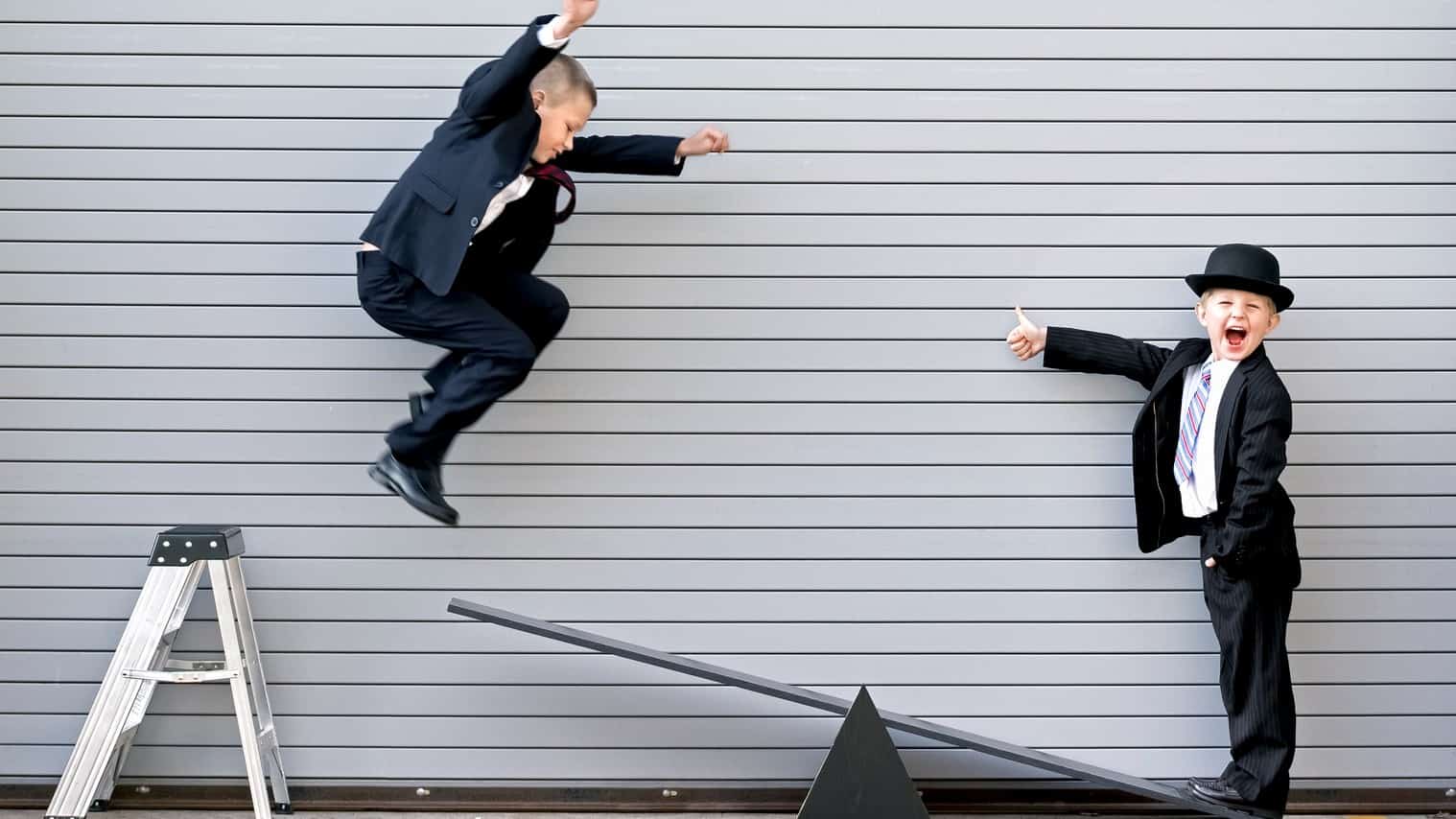 one young boy jumps off a step ladder and is captured mid-air about to land on a seesaw where his friend is standing with a wide smile on his face looking at the camera and holding his thumbs up as though he is excited for the ride to come. Both young boys are wearing business suits.