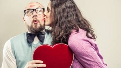 a geeky looking man wearing a vest and a bow tie clutches a stuffed love heart as he is covered in lipstick kisses from an attractive woman leaning into him and kissing him on the cheek.