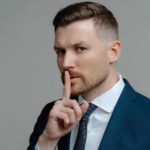 A male broker wearing a dark blue suit and tie puts his finger to his lips to signal a secret tip about the Xero share price