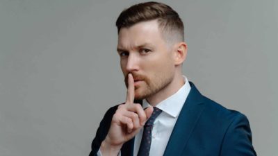 A male broker wearing a dark blue suit and tie puts his finger to his lips to signal a secret tip about the Xero share price