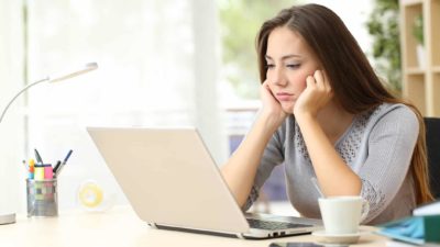 a young woman sits with her hands holding up her face as she stares unhappily at a laptop computer screen as if she is disappointed with something she is seeing there.