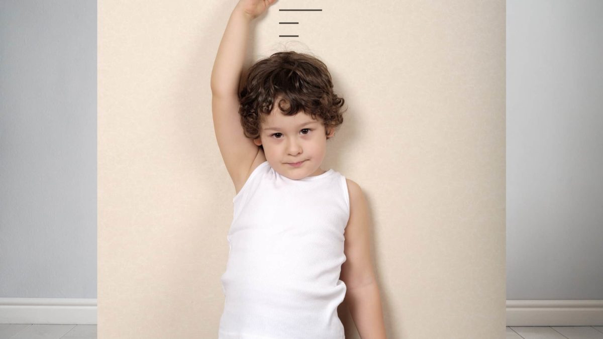 A young child stands against a wall holding measuring tape behind them as they wish not to be so short