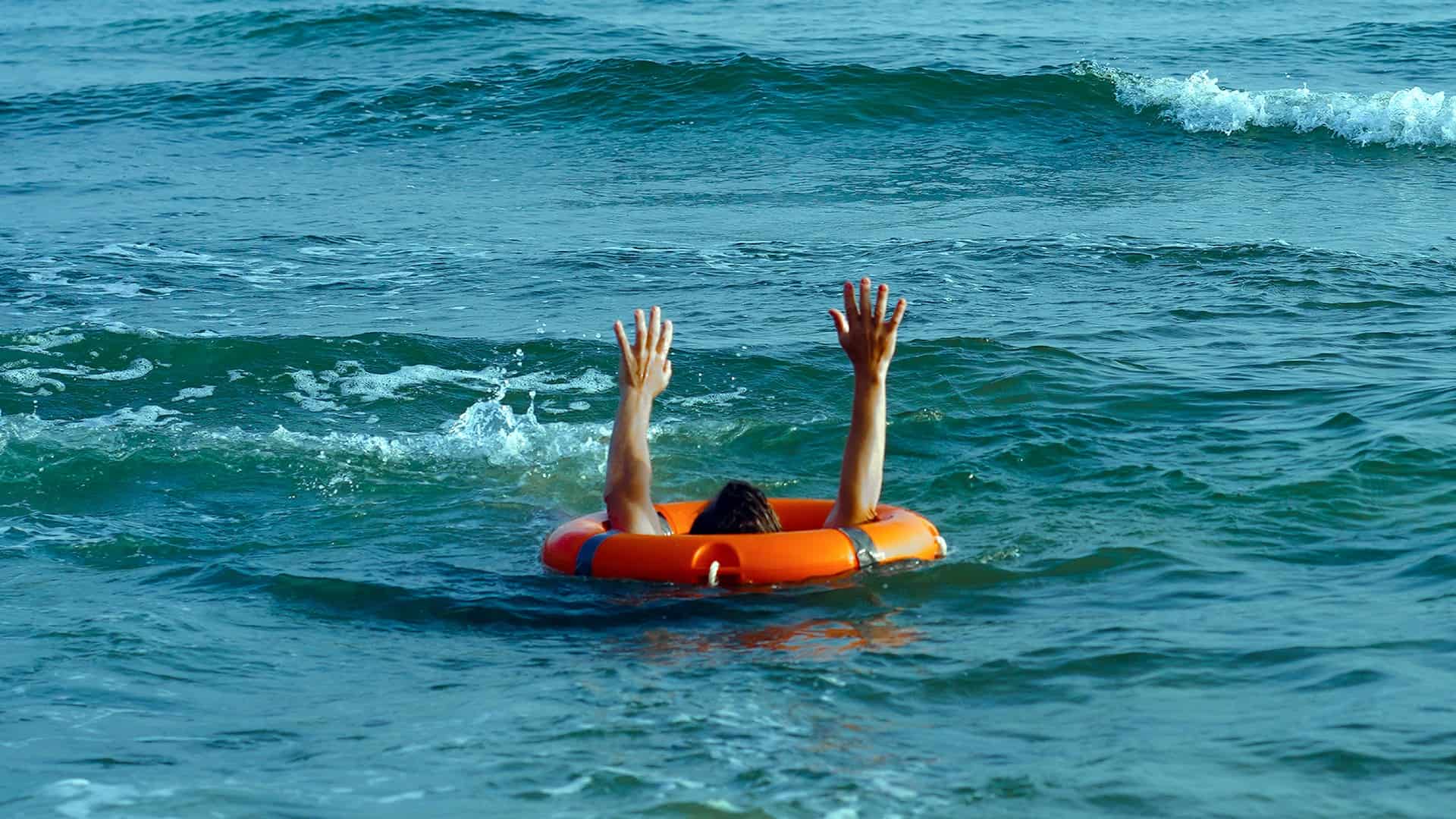 a person holds their hands up through the middle of a rubber lifesaving ring while swimming in relatively calm conditions at a beach.