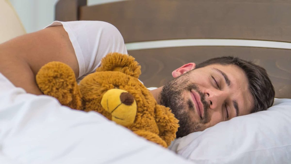 A man sleeps in a bed with white sheets while holding a teddy bear and a smile on his face.