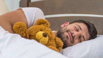 A man sleeps in a bed with white sheets while holding a teddy bear and a smile on his face.