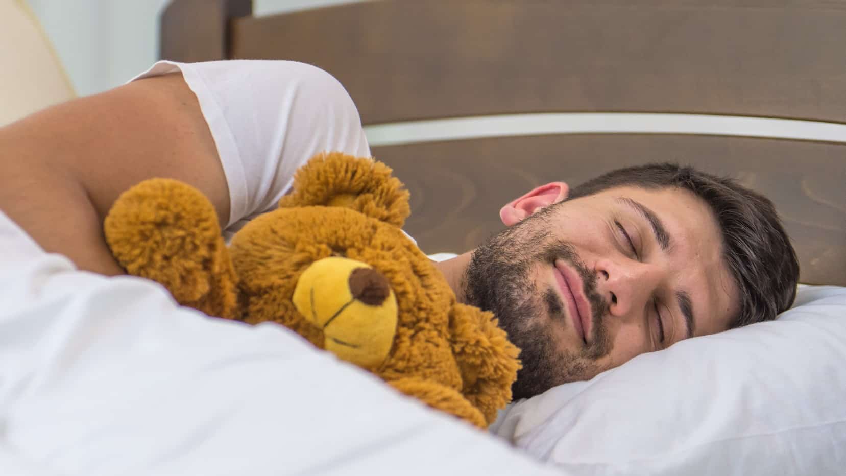 A man sleeps in a bed with white sheets while holding a teddy bear representing the improving performance of the Adairs share price