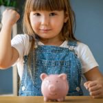 Small girl giving a fist bump with a piggy bank in front of her.