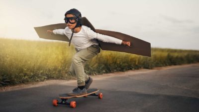 Kid on a skateboard with cardboard wings soars along the road.