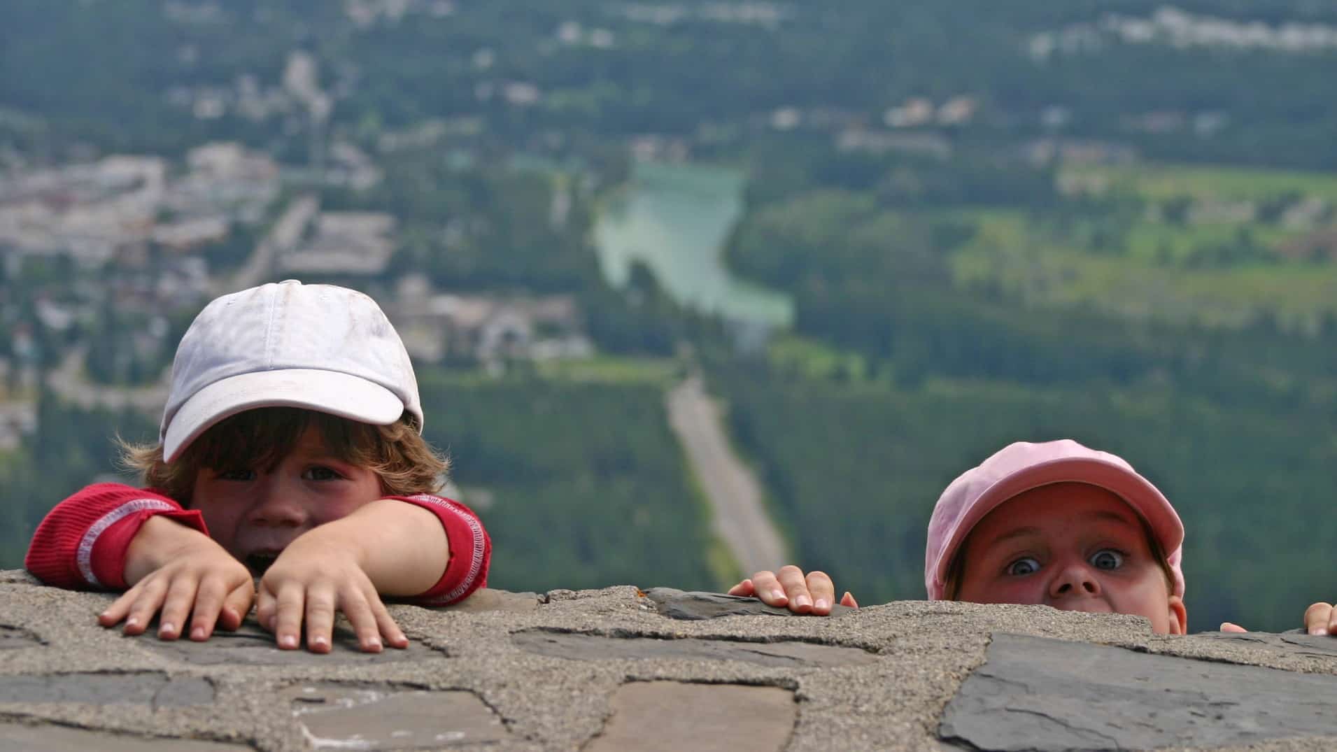 two young children wearing caps poke their heads above a all with a panoramic view of a lush countryside behind them.