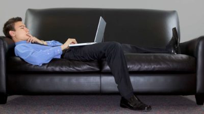 a man wearing a business shirt and pants reclines on a leather sofa with his laptop computer resting on his stomach as he looks concerned at what he's reading on the screen.