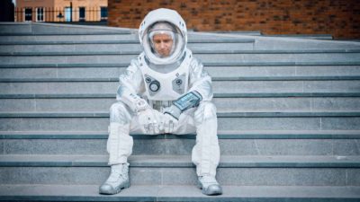 a man in full astronaut suit sits forlornly on a set of concrete steps with a sorrowful look on his face beneath his rounded space helmet.