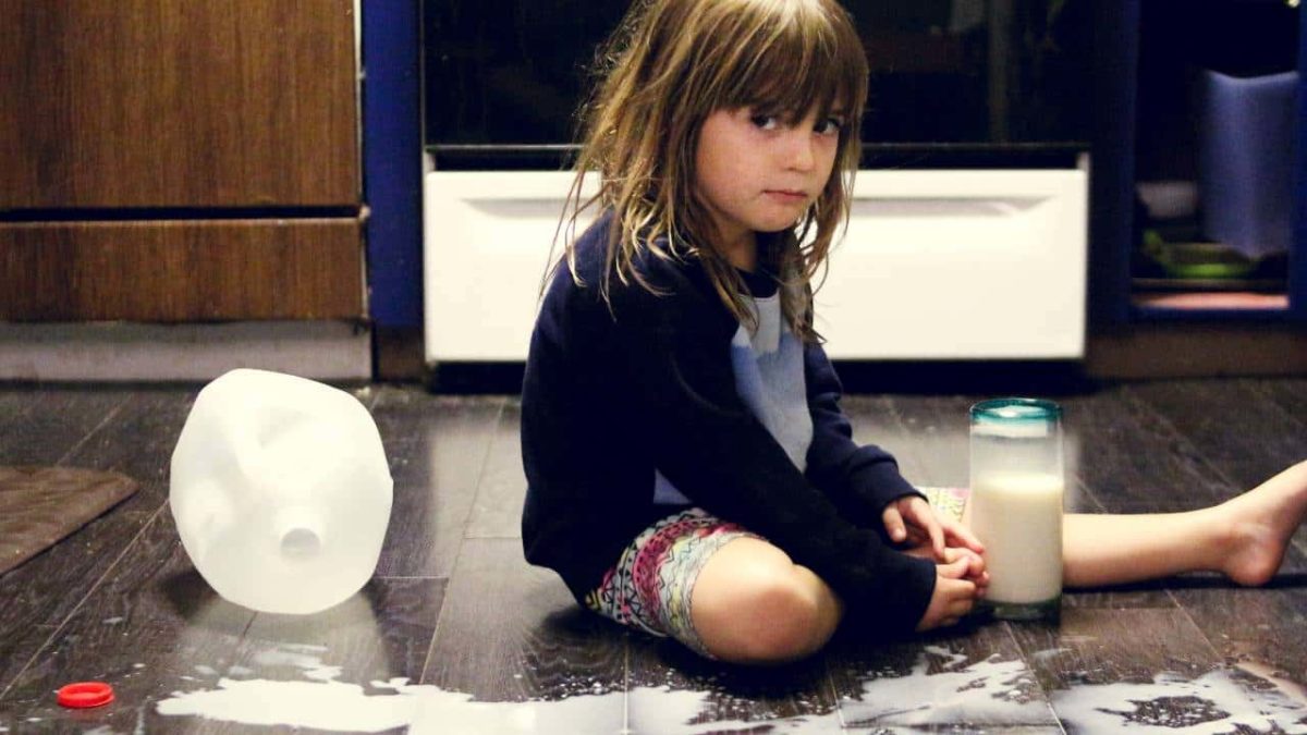 A young girl sits on her kitchen floor holding a glass of milk with an empty A2 Milk Company bottle next to her and milk puddles on the floor