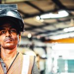 A steel worker peers out from under his protective headwear which is tipped back on his head as he stares solemnly straight ahead with steel production equipment in the background.
