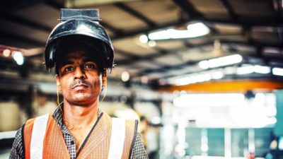 A steel worker peers out from under his protective headwear which is tipped back on his head as he stares solemnly straight ahead with steel production equipment in the background.