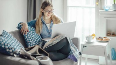 A young woman sits on a sofa in a stylish home with her laptop computer balanced on her knee and smiles with a satisfied look on her face at what she's seeing on the screen.