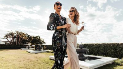Two stylish female friends stroll along a grassy area holding champagne glasses and laughing as they enjoy the good life due to Pilbara Minerals share price gain in recent times