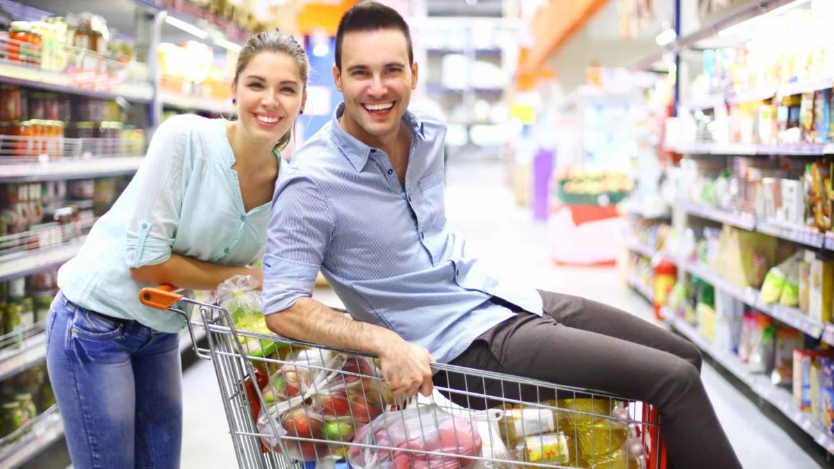 Happy man on a supermarket trolley full of groceries with a woman standing beside him.
