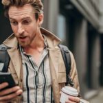 A cool young man walking in a laneway holding a takeaway coffee in one hand and his phone in the other reacts with surprise as he reads the latest news on his mobile phone