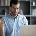 A surprised man sits at his desk in his study staring at his computer screen with his hands up.