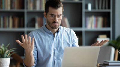 A surprised man sits at his desk in his study staring at his computer screen with his hands up.